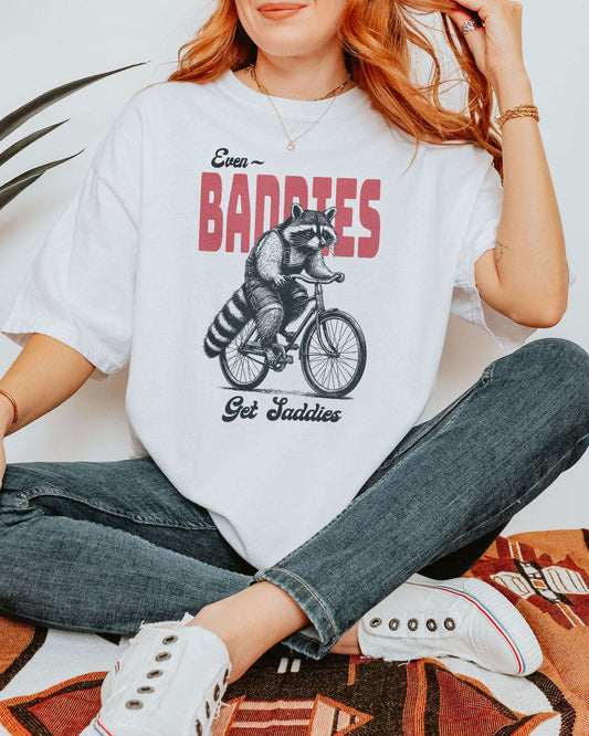 Woman wearing a white "Even Baddies Get Saddies" shirt with a funny retro graphic of a raccoon riding a bicycle.