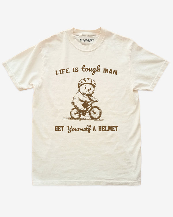 'Life Is Tough Man Get Yourself A Helmet' shirt in ivory with a retro graphic of a little bear riding a bycicle.