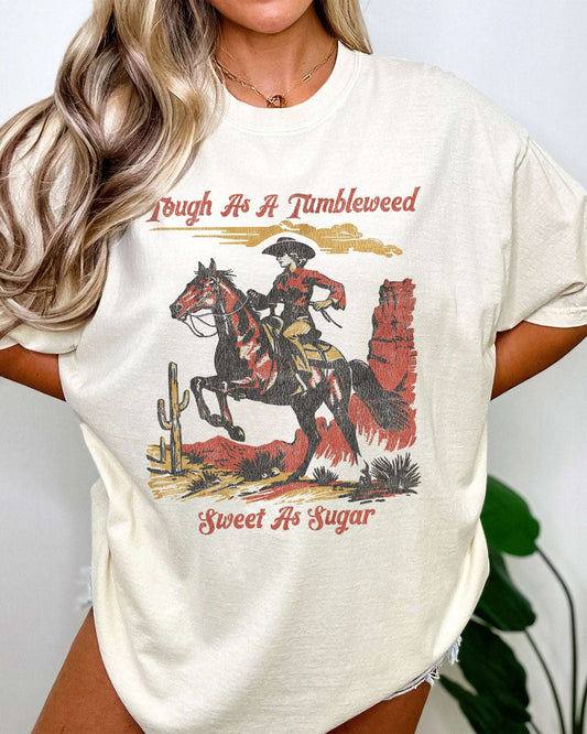 Woman wearing ivory "Tough As A Tumbleweed Sweet As Sugar" shirt with retro graphic of cowgirl riding horse through desert