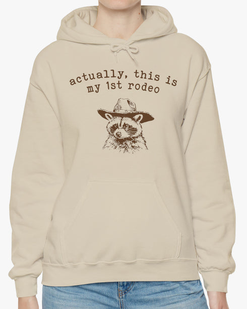 Woman wearing an 'Actually This is My First Rodeo' hoodie in sand beige, featuring a retro western graphic design and unisex fit.