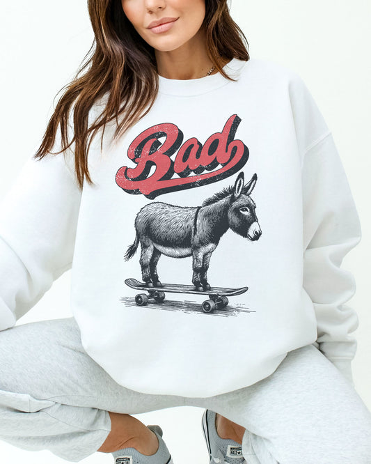Woman wearing a white sweatshirt with a retro graphic of a donkey riding a skateboard and the word 'Bad' above it.