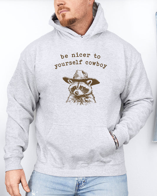 Man wearing an ash gray hoodie featuring a funny retro cowboy raccoon graphic and the quote 'Be Nicer To Yourself Cowboy'.