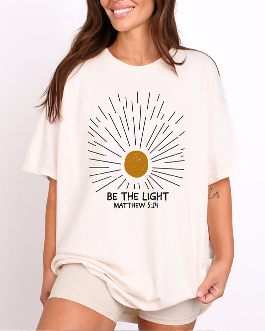 Model wearing natural color shirt with 'Be The Light - Matthew 5:14' graphic design