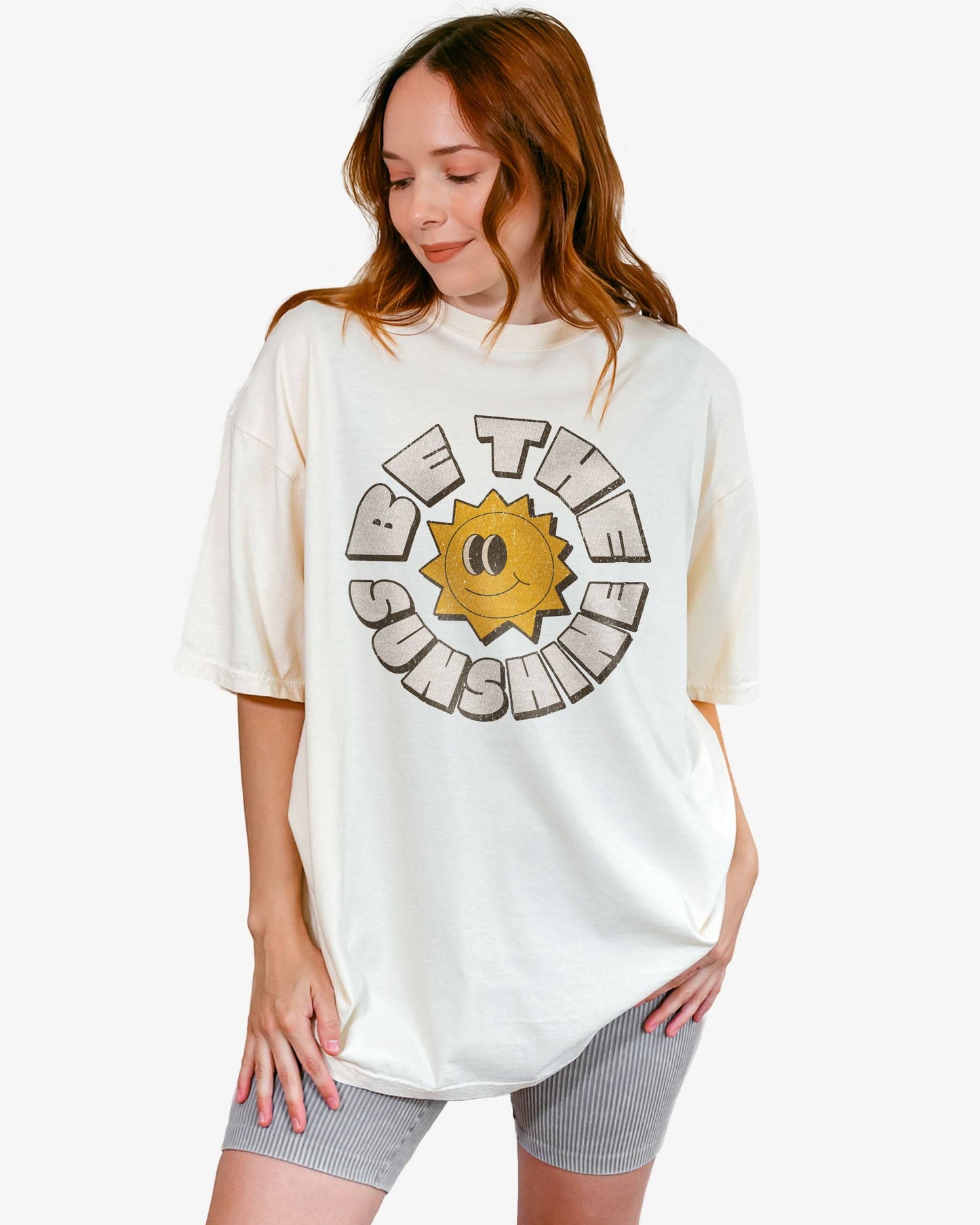 Woman wearing an ivory Be The Sunshine shirt with a retro boho sun graphic.