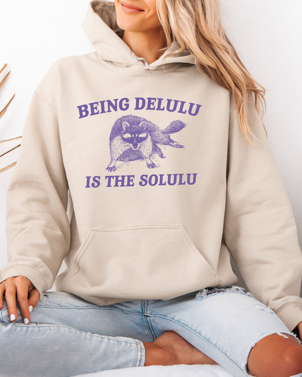Woman wearing a sand beige hoodie featuring a unny retro-style fat raccoon graphic and the quote 'Being Delulu Is The Solulu'.