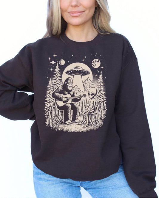 Woman wearing a dark chocolate color sweatshirt featuring a retro graphic of Bigfoot playing guitar with an alien and UFO.
