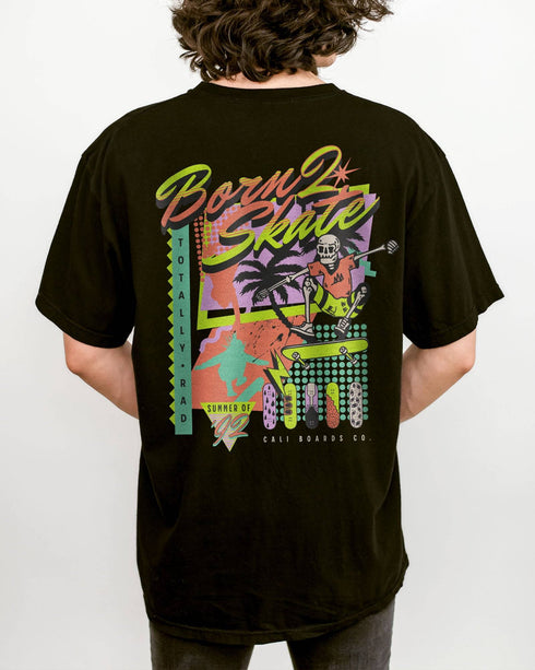 Man wearing a black “Born 2 Skate” shirt with a retro 90s-style graphic of a skeleton skateboarding and bold skateboard designs.