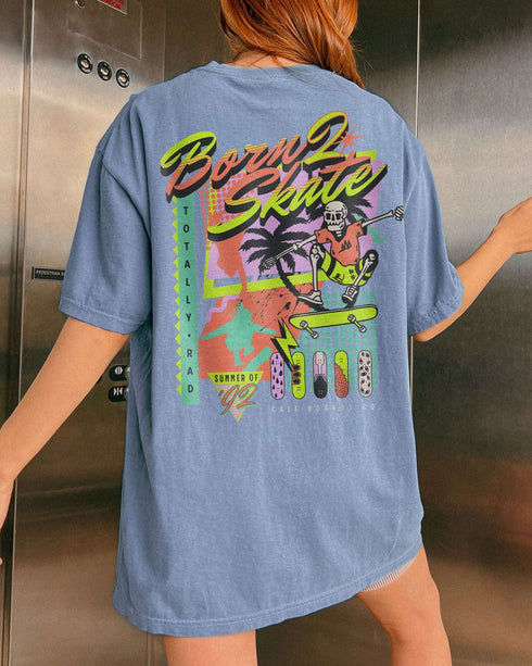 Woman wearing a blue jean “Born 2 Skate” shirt with a retro 90s-style graphic of a skeleton skateboarding and bold skateboard designs.