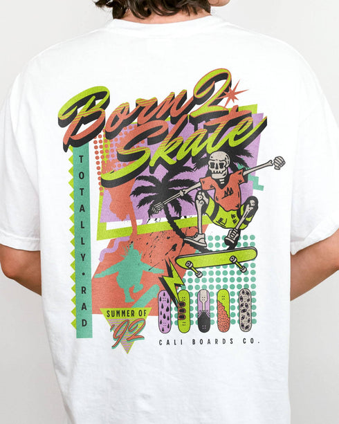 Close-up of a man wearing a white “Born 2 Skate” shirt with a retro 90s-style graphic of a skeleton skateboarding and bold skateboard designs.