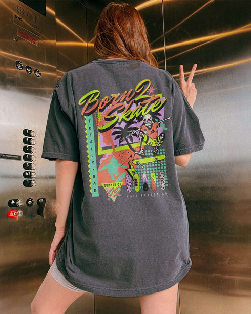 Woman wearing a pepper “Born 2 Skate” shirt with a retro 90s-style graphic of a skeleton skateboarding and bold skateboard designs.