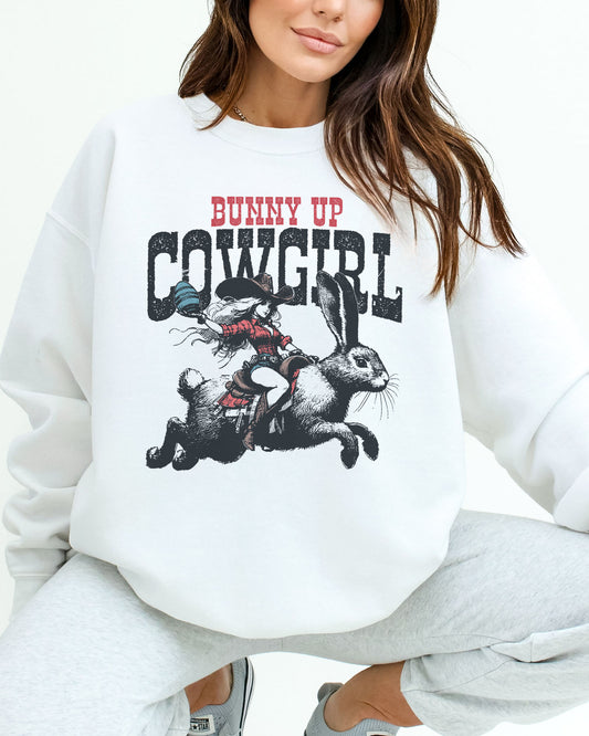 Woman wearing a white sweatshirt featuring a funny retro graphic of a cowgirl riding an easter bunny and the quote 'Bunny Up Cowgirl'.