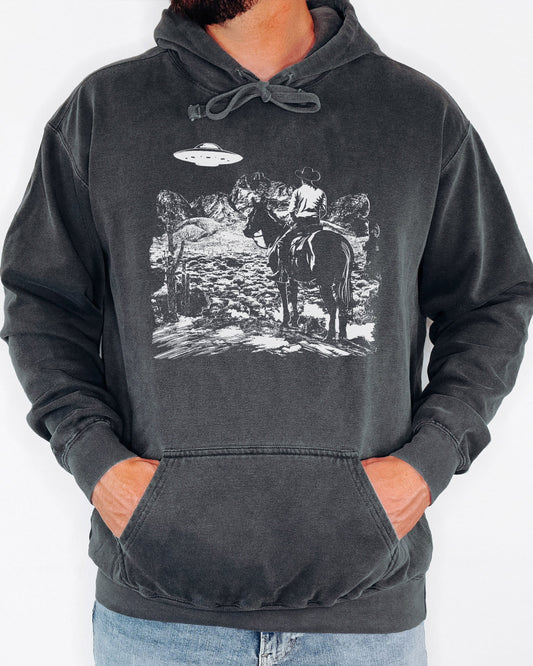 Man wearing a pepper-colored "Cowboy UFO Encounter" hoodie with a retro graphic of a cowboy riding through a desert as a UFO hovers above.