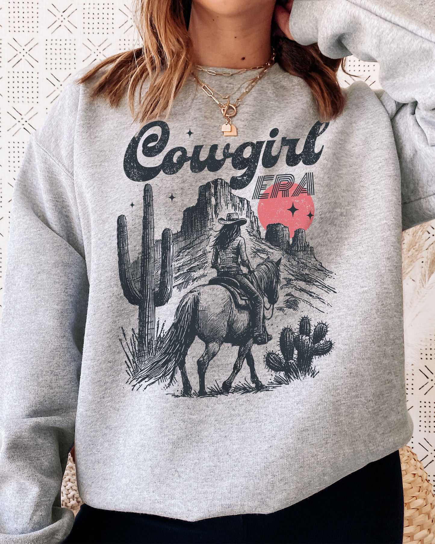 Woman wearing an ash gray sweatshirt featuring a retro cowgirl riding a horse through a Western desert with the lettering 'Cowgirl Era'.