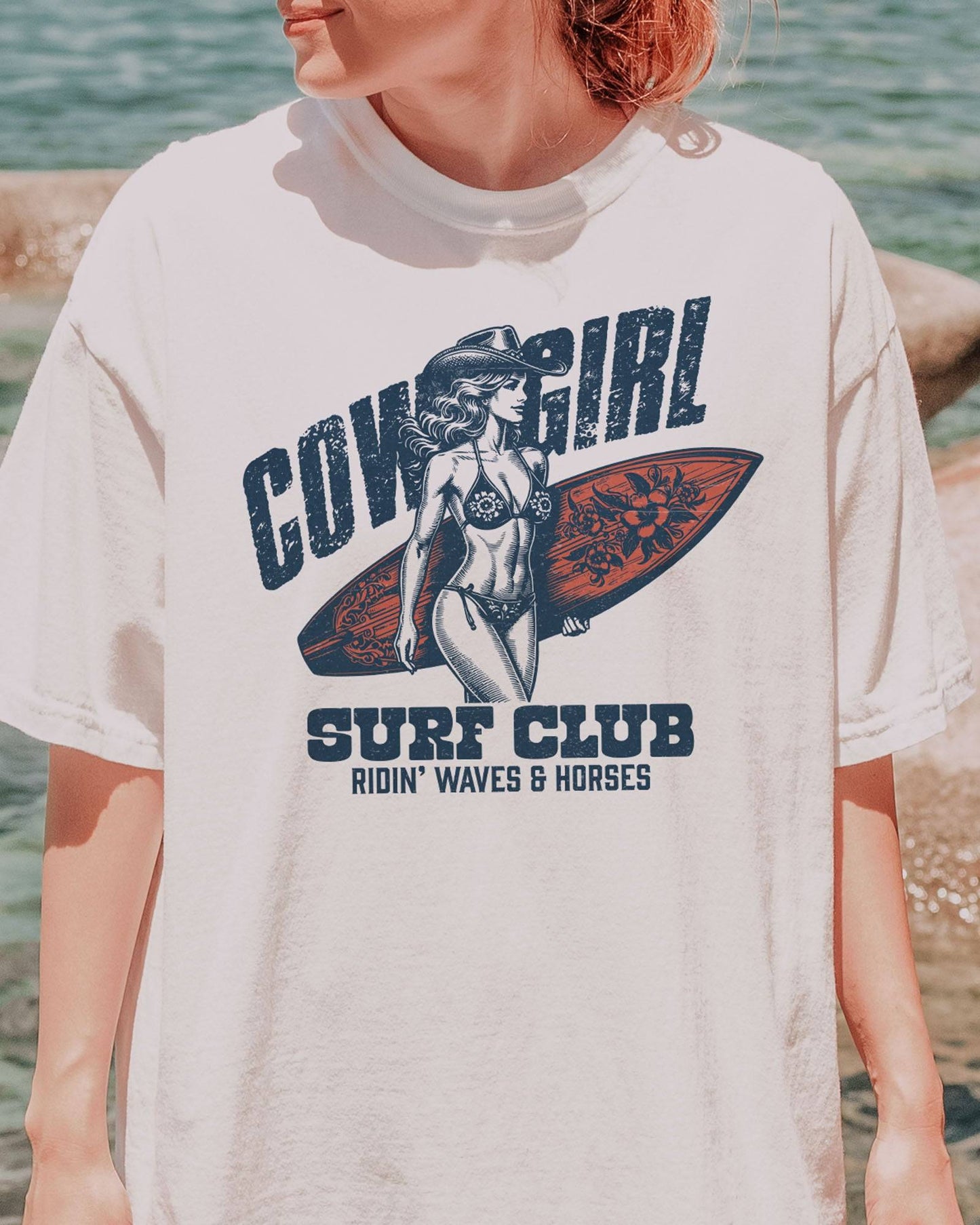 Close-up of woman wearing a white graphic tee at the beach with the quote "Cowgirl Surf Club" and a retro graphic of a cowgirl in a bikini with a surfboard in her hand.