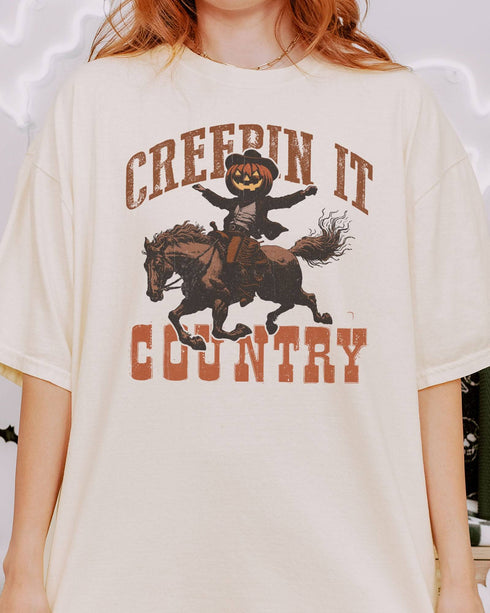 Close-up of woman wearing an ivory "Creepin It Country" shirt with a retro Halloween graphic of a pumpkin-headed cowboy riding a horse.