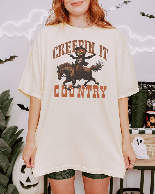 Woman wearing an ivory "Creepin It Country" shirt with a retro Halloween graphic of a pumpkin-headed cowboy riding a horse.