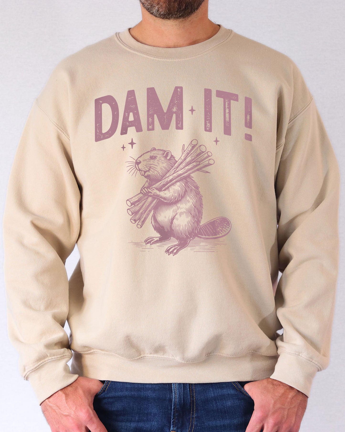 Man wearing a sand beige “Dam It” sweatshirt with a retro-style beaver graphic and funny expression.