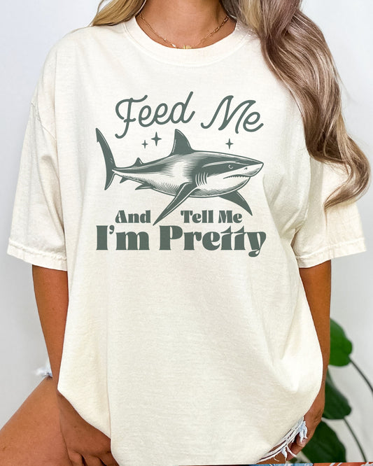 Woman wearing a funny "Feed Me And Tell Me I'm Pretty" shirt in natural color with a retro shark graphic on the front of the tee