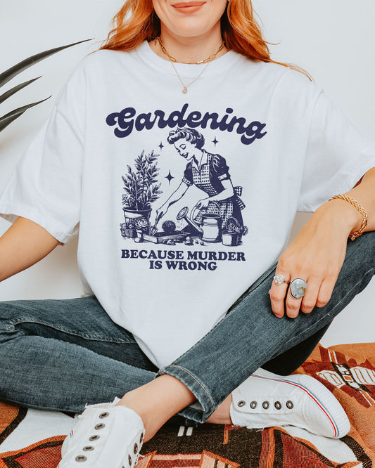 Woman wearing a white "Gardening Because Murder Is Wrong" shirt with a funny retro graphic of a housewife gardening in a flower bed.