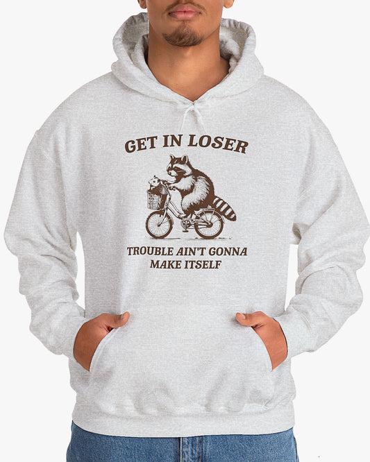 Man wearing ash gray color hoodie with the quote 'Get In Loser, Trouble Ain't Gonna Make Itself' and a funny retro raccoon riding a bycicle graphic