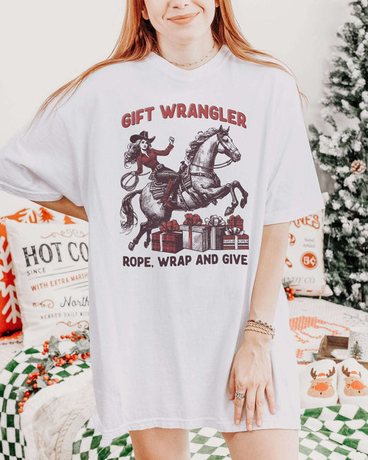 Woman wearing a white "Gift Wrangler – Rope, Wrap and Give" shirt with a funny retro cowgirl Christmas graphic.