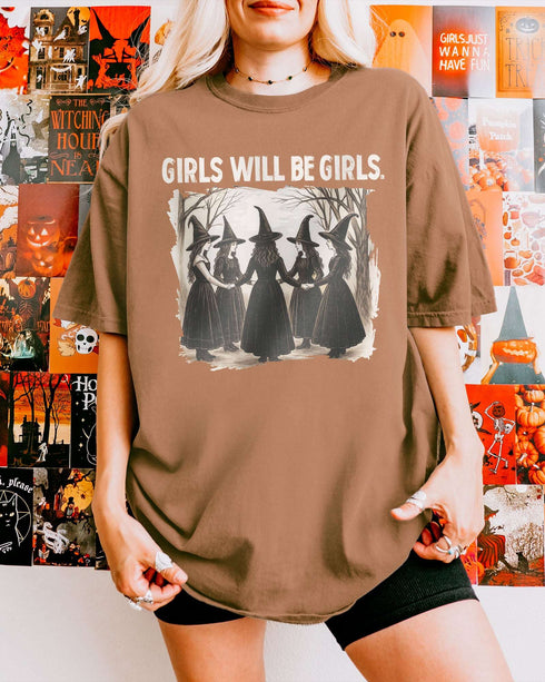 Woman wearing an espresso-colored “Girls Will Be Girls” shirt with a retro graphic of witches performing a witchy ritual.