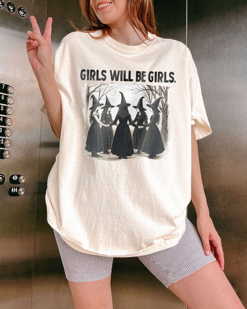 Woman wearing an ivory-colored “Girls Will Be Girls” shirt with a retro graphic of witches performing a witchy ritual.