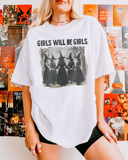Woman wearing a white-colored “Girls Will Be Girls” shirt with a retro graphic of witches performing a witchy ritual.