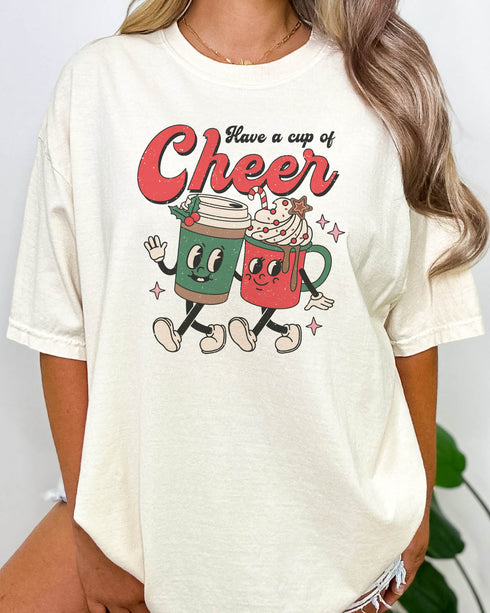 Woman wearing a retro Christmas coffee graphic shirt in natural beige color with the quote "Have A Cup of Cheer"