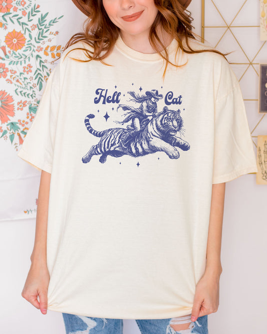 Woman wearing an ivory "Hell Cat" shirt with a retro graphic of a cowgirl riding a tiger.