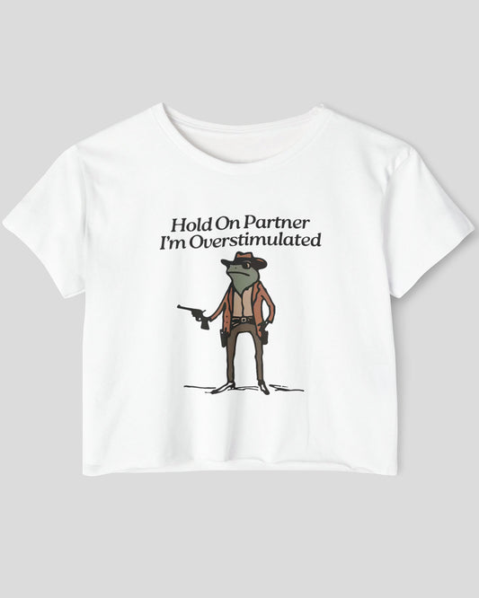 Flat lay white 'Hold On Partner, I'm Overstimulated' crop top featuring a funny retro cowboy frog graphic.