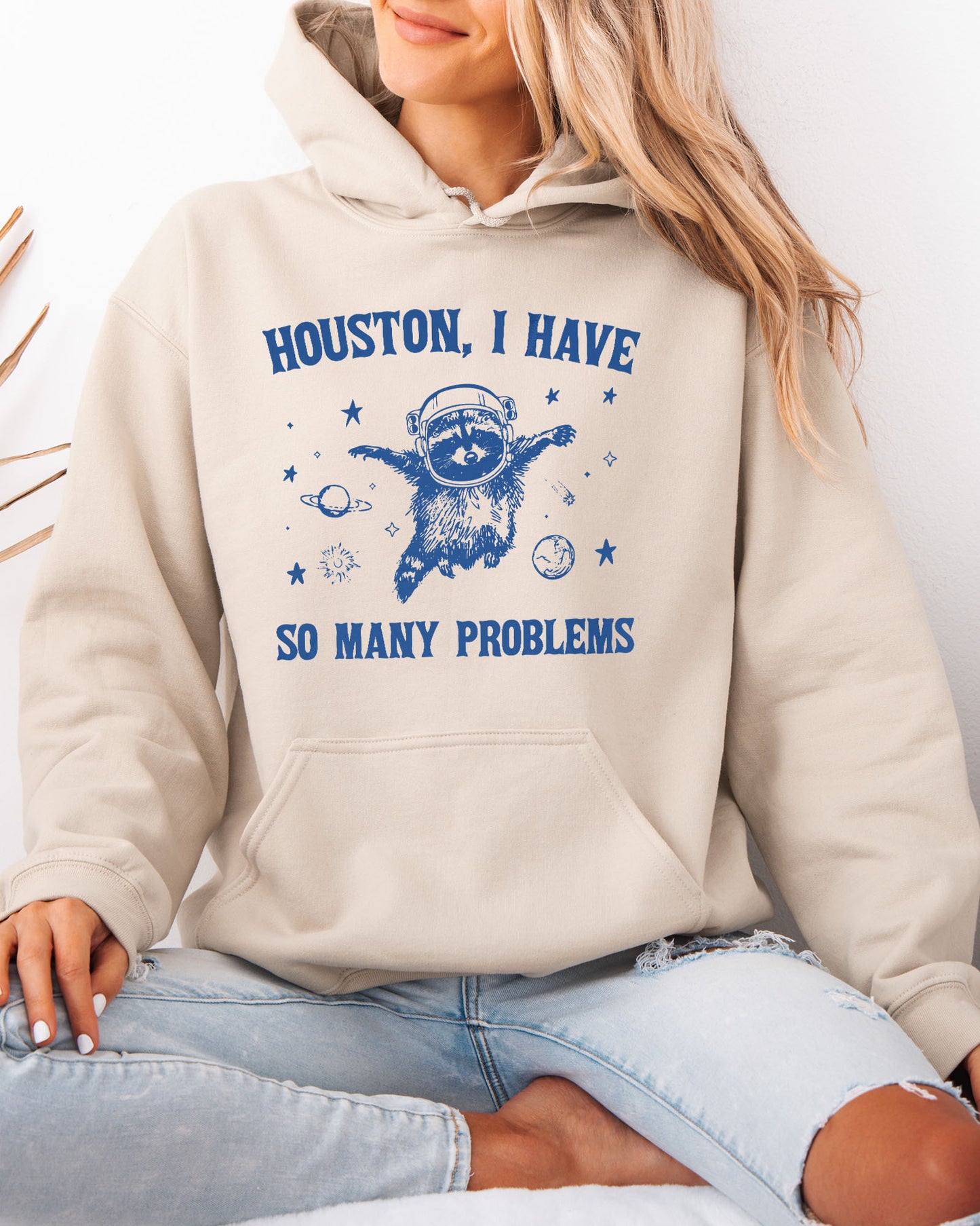 Woman wearing a sand beige hoodie featuring a funny retro-style astronaut raccoon space graphic and the quote 'Houston I Have So Many Problems'.