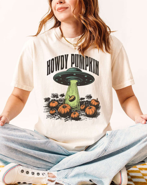 Woman wearing an ivory "Howdy Pumpkin" shirt with a retro graphic of a UFO abducting pumpkins from a field.