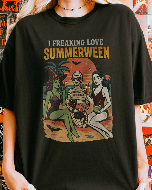 Close-up of woman wearing a black "I Freaking Love Summerween" shirt with a retro graphic of two witches and a mummy relaxing on the beach.