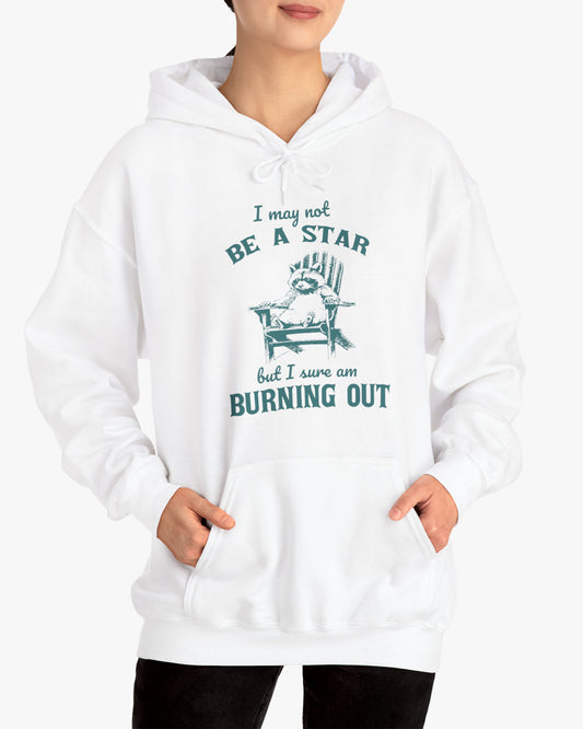 Woman wearing a 'I May Not Be A Star But I Sure Am Burning Out' hoodie in white, featuring a retro raccoon graphic design and unisex fit.