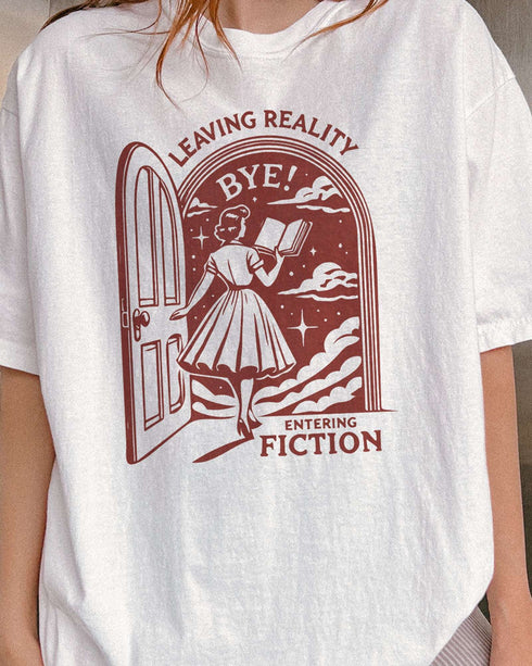 Close-up of woman wearing a white 'Leaving Reality Entering Fiction' shirt with a funny retro graphic of a woman reading while stepping through a door into a new universe.