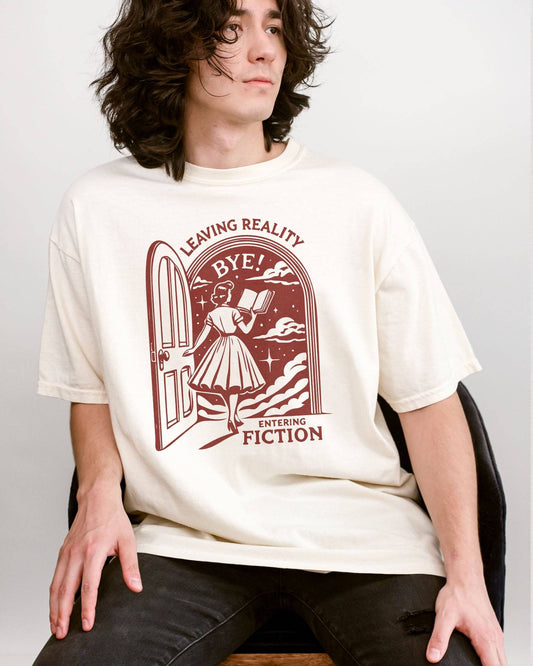 Man wearing an ivory 'Leaving Reality Entering Fiction' shirt with a funny retro graphic of a woman reading while stepping through a door into a new universe.
