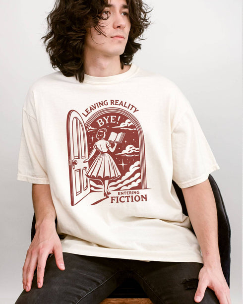 Man wearing an ivory 'Leaving Reality Entering Fiction' shirt with a funny retro graphic of a woman reading while stepping through a door into a new universe.