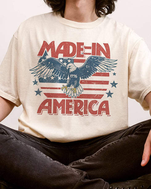 Close-up of man wearing "Made In America 1776" shirt in ivory color with retro graphic of a hawk in USA red-white-and-blue colors.
