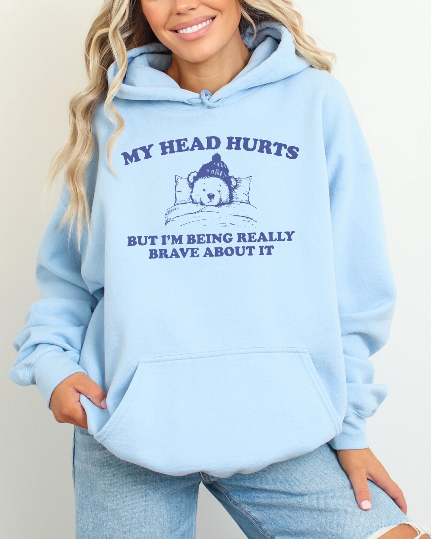 Woman wearing a light blue hoodie featuring a funny retro-style bear lying in bed with the quote 'My Head Hurts But I'm Being Really Brave About It'.