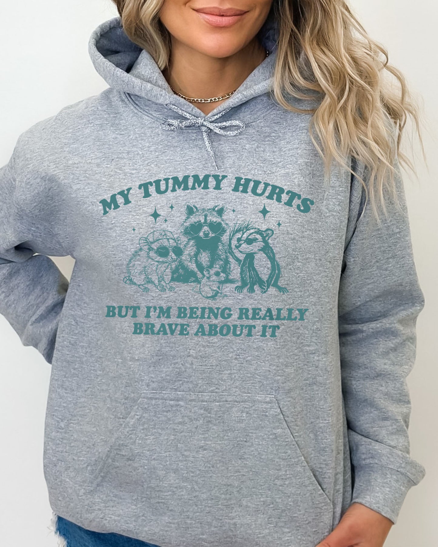 Woman wearing a sport gray hoodie with a retro raccoon gang graphic and the quote 'My Tummy Hurts But I'm Being Really Brave About It'.