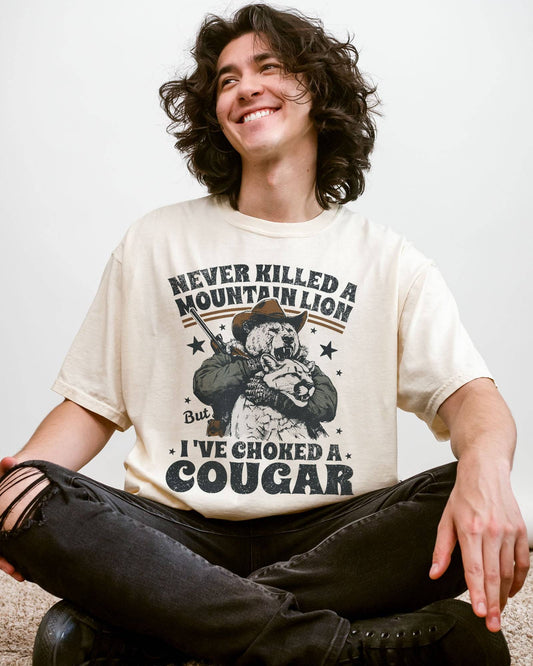 Man wearing an ivory "Never Killed a Mountain Lion But I've Choked a Cougar" shirt with a funny retro graphic of a bear choking a cougar.