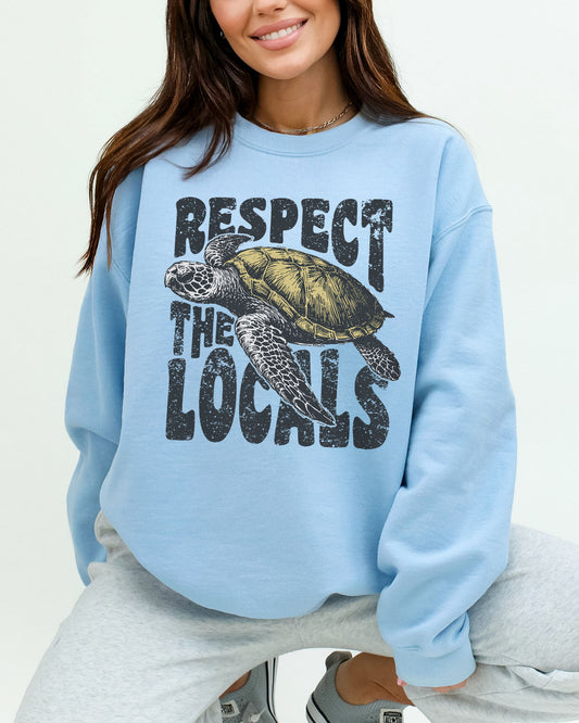 Woman wearing a light blue sweatshirt featuring a retro sea turtle graphic and the quote 'Respect The Locals'.