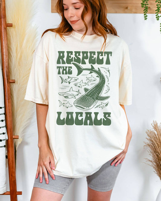 Woman wearing an ivory-colored ‘Respect The Locals’ shirt with a retro graphic of whales, sharks, and fish.