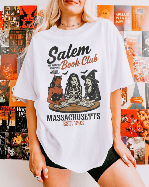 Woman wearing a white 'Salem Book Club Massachusetts 1692' shirt with a retro Halloween graphic of a devil, skeleton, and witch sitting together reading at a table.
