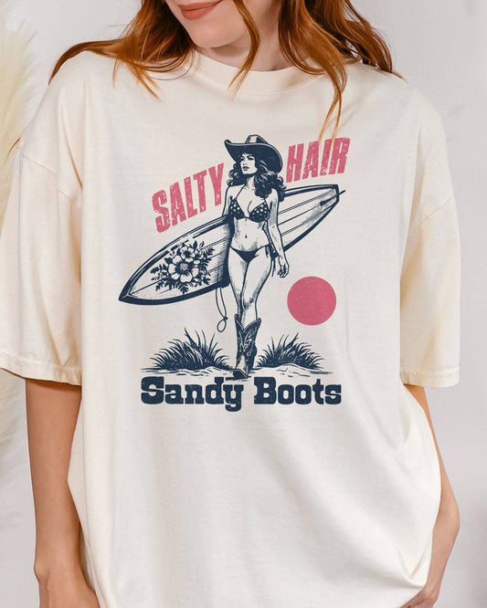 Close-up of woman wearing "Salty Hair Sandy Boots" shirt in white color with retro graphic of a cowgirl with a surfboard under her arm.