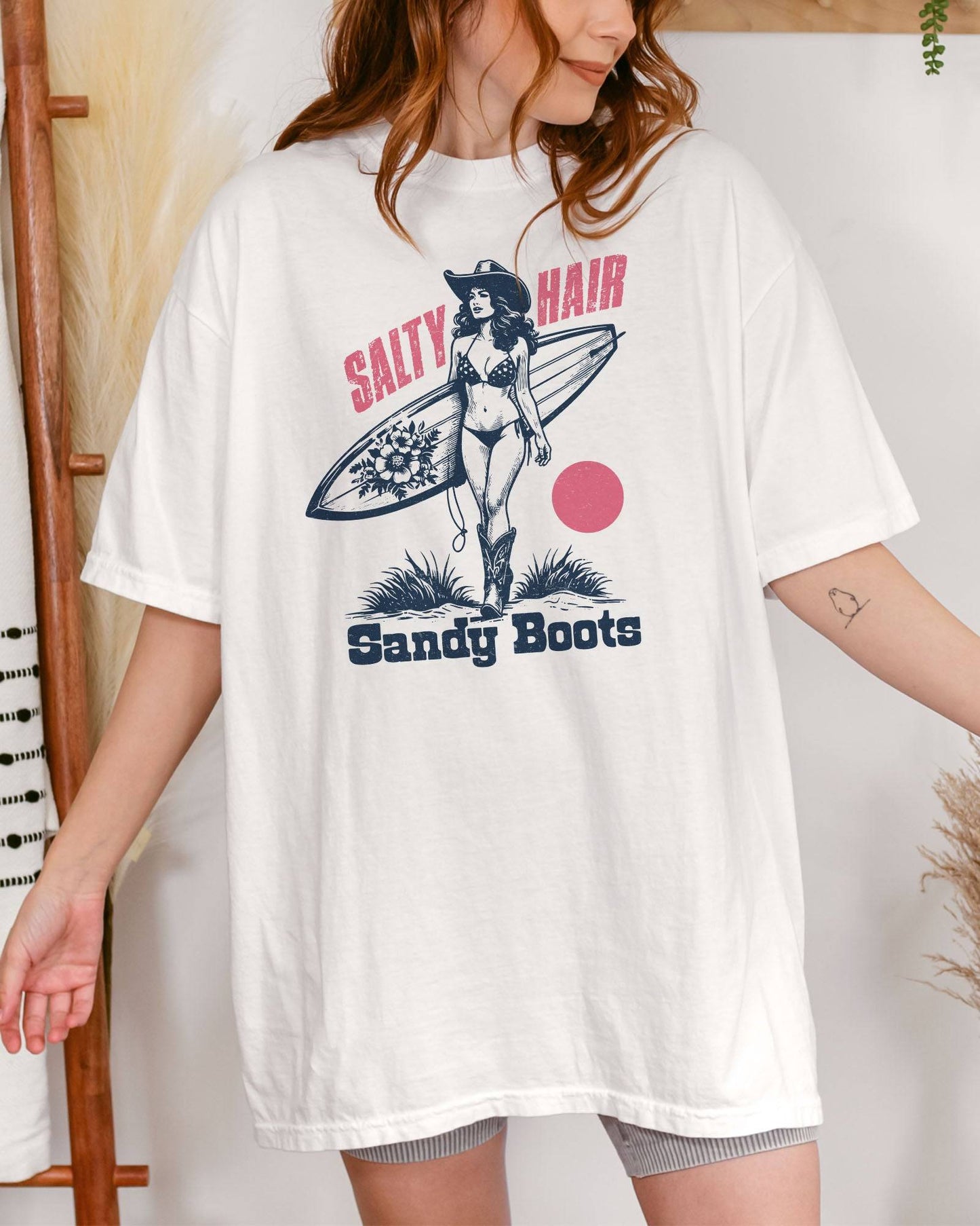 Woman wearing "Salty Hair Sandy Boots" shirt in white color with retro graphic of a cowgirl with a surfboard under her arm.