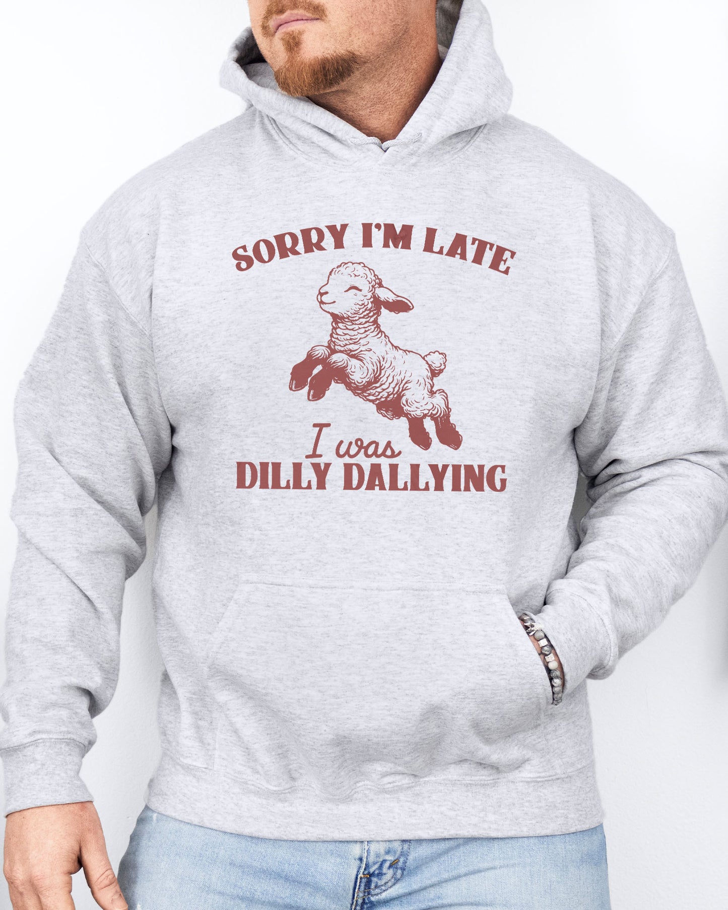 Man wearing an ash gray hoodie featuring a funny retro-style baby sheep graphic and the quote 'Sorry I'm Late, I Was Dilly Dallying '.