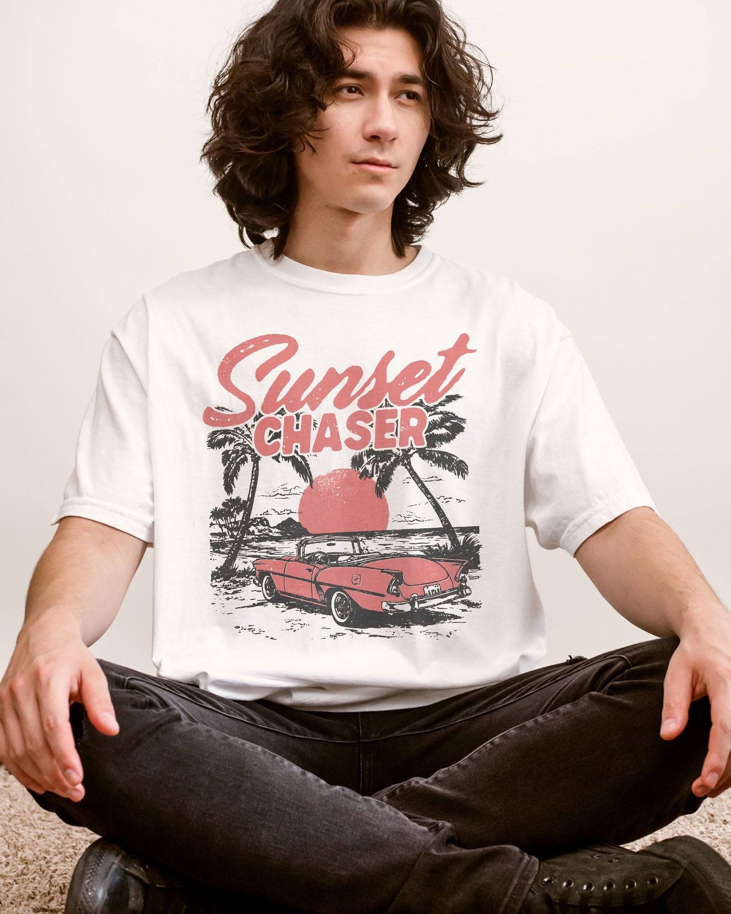 Man wearing white shirt with the quote "Sunset Chaser" and a retro car on a beach scenery graphic.