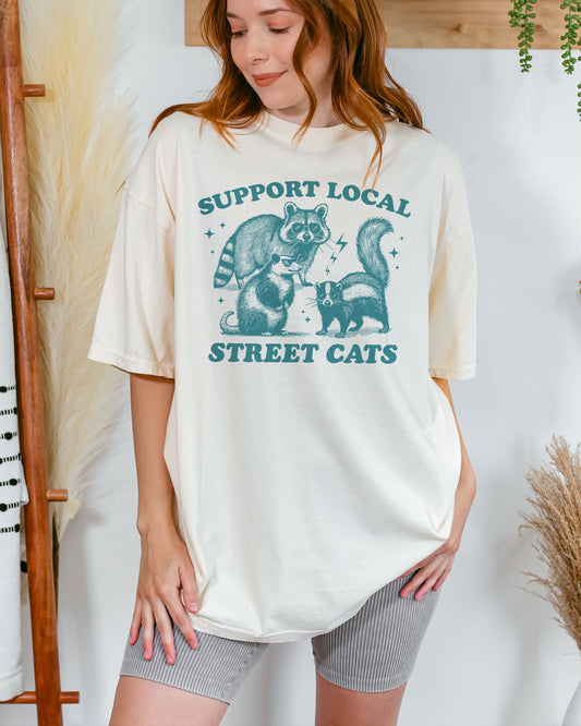 Woman wearing an ivory "Support Local Street Cats" shirt with a retro graphic of a raccoon, opossum, and skunk wearing sunglasses.